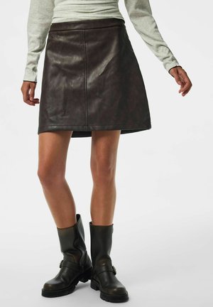 Personne portant une jupe en cuir marron foncé avec un haut léger à manches longues et des bottes en cuir noires mi-mollet sur fond blanc.