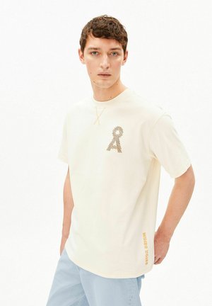 Jonge man met kort donker haar, gekleed in een crème T-shirt met een gestructureerd "A"-logo op de borst en lichtblauwe jeans, staand met één hand achter de rug.