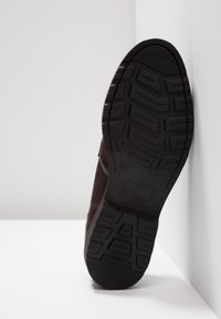 Semelle noire d’une seule botte marron foncé appuyée contre un mur blanc, montrant un motif de bande de roulement et la taille 37 indiquée.