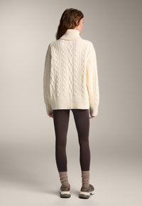 Dikke crèmekleurige kabelgebreide trui met een hoge kraag, gecombineerd met bruine leggings en enkelkousen. Het ontwerp heeft gestructureerde diamantpatronen.