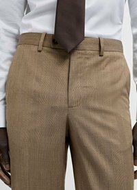 Pantaloni în carouri maro, cu un finisaj texturat și o croială ajustată, având buzunare laterale și un brâu cu bucle pentru curea, asortați cu o cămașă albă și o cravată maro.