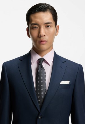 Veste de costume bleu marine à finition texturée, chemise rose pâle, et cravate noire avec petits motifs carrés blancs. Mouchoir de poche blanc visible.