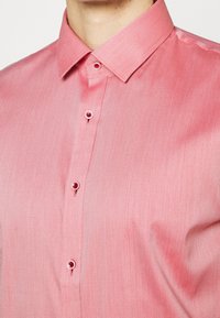 Uomo che indossa una camicia rosa con bottoni e colletto a punta, che mostra la parte superiore del petto e del collo.