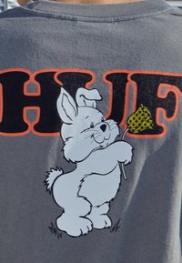 Graues T-Shirt mit einem großen Grafikmotiv eines weißen Cartoon-Hasen, der eine gelbpunktierte Flagge hält, mit dem fetten "HUF"-Text darüber in Orange.