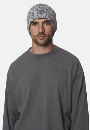 Hombre con barba corta que lleva un gorro gris con patrón y una sudadera holgada de color gris oscuro contra un fondo claro y liso.