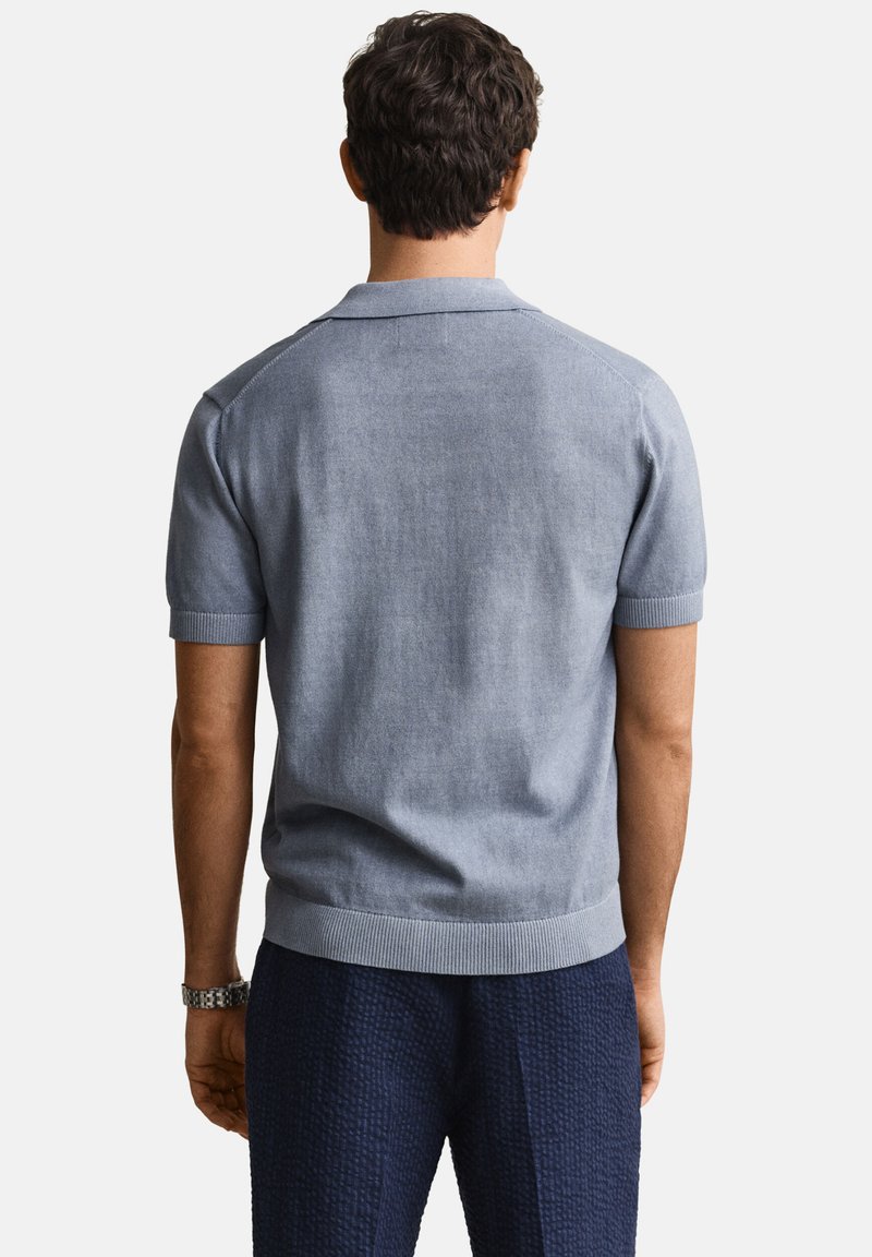 Hombre de cabello oscuro de espaldas, vestido con una camisa polo tejida de manga corta azul claro y pantalones texturizados azul marino sobre un fondo blanco.