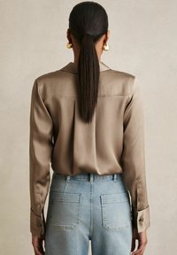 Femme avec une longue queue de cheval foncée portant des boucles d’oreilles anneaux dorées, un chemisier en satin taupe et un jean taille haute bleu clair, vue de dos.