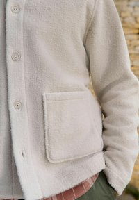 White fleece jacket with a button-up front, textured surface, and a patch pocket. Worn over a plaid shirt, featuring long sleeves.
