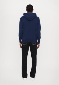Marineblauer Hoodie mit Kordelzugkapuze, langen Ärmeln und gerippten Bündchen. Kombiniert mit schwarzer Hose und weißen Turnschuhen, von hinten gezeigt.
