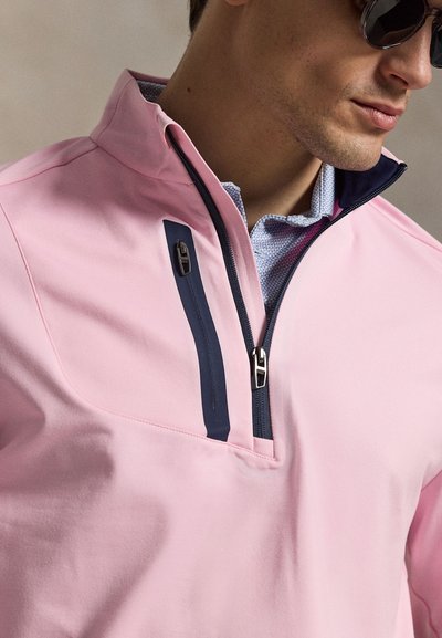Άνδρας που φοράει ανοιχτό ροζ μπουφάν με φερμουάρ μέχρι το ύψος του στήθους, με σκούρο μπλε φερμουάρ και λεπτομέρειες στην τσέπη πάνω από πουκάμισο με πατρόν και σκούρα γυαλιά ηλίου.