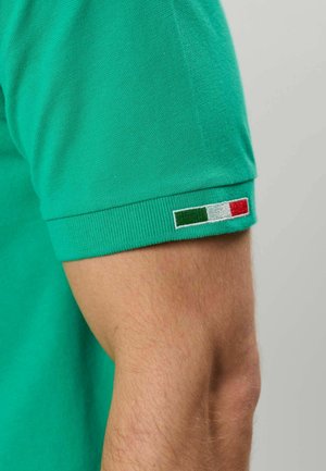 Arm trägt ein grünes kurzärmeliges Hemd mit einem kleinen italienischen Flaggen-Patch am Ärmelsaum, sichtbar vor schlichtem Hintergrund.