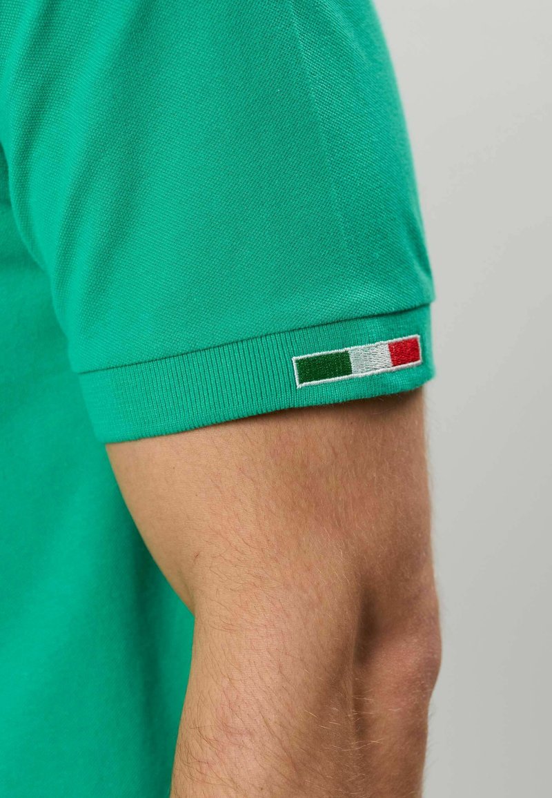 Bras portant une chemise verte à manches courtes avec un petit écusson du drapeau italien sur le poignet visible sur un fond uni.