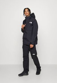 Giacca impermeabile nera con cappuccio e tasche anteriori, abbinata a pantaloni coordinati. Entrambi presentano un logo bianco sul petto e sulla coscia.