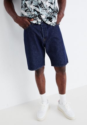 Shorts in denim scuro con vestibilità rilassata, dotati di cinque tasche e gamba dritta. Indossati con una camicia a fantasia e sneakers bianche.