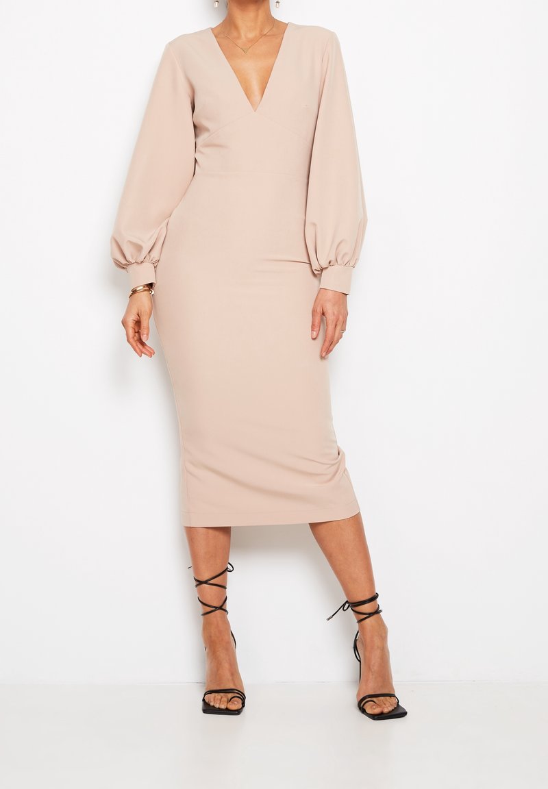 Femme portant une robe midi beige avec des manches longues bouffantes et un profond décolleté en V, associée à des sandales noires à talons hauts à brides.