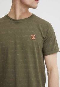 Das olivgrüne T-Shirt hat einen Rundhalsausschnitt und eine subtile Textur mit roten und weißen Einsprenkelungen. Ein kleines oranges Logo befindet sich auf der linken Brustseite.
