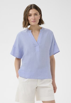 Vrouw met schouderlang bruin haar die een lichtblauw T-shirt met korte mouwen en V-hals en witte korte broek draagt, staande met handen in de zakken.