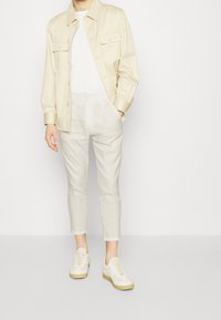 Osoba v béžové košilové tričko přes bílé tričko, béžových zkrácených kalhotách a béžových teniskách, stojící na bílém pozadí.