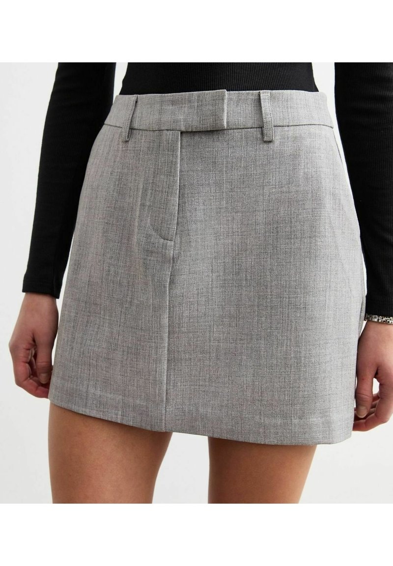 Femme portant une mini-jupe grise taille haute avec passants pour ceinture et fermeture frontale cachée, associée à un top noir à manches longues.