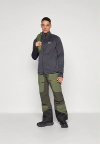 Herreutendørsantrekk med en mørkegrå jakke med glidelås og grønne bukser med flere paneler og cargolommer, med svarte detaljer og praktiske lommer.