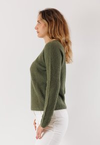 Donna con capelli lunghi e ondulati, che indossa un maglione lavorato a maglia verde e pantaloni bianchi, in piedi di profilo contro uno sfondo chiaro e uniforme.
