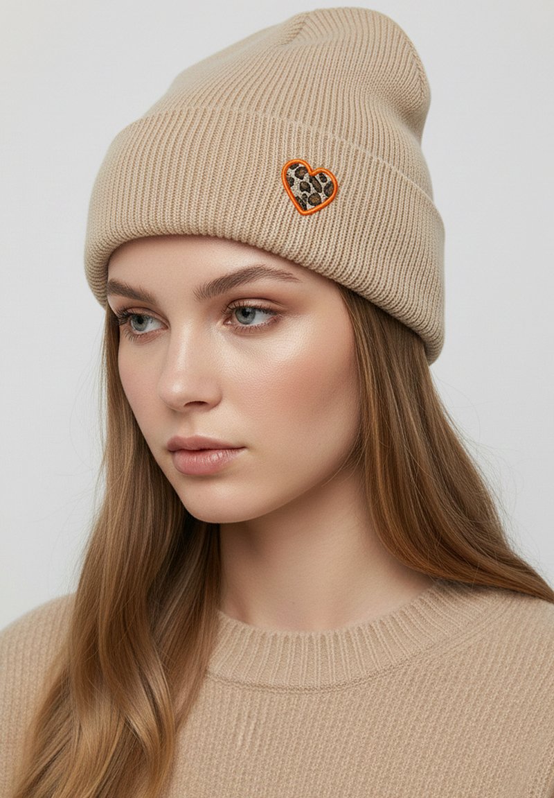 Beige knitted beanie with ribbed texture, featuring a heart-shaped leopard print patch outlined in orange.