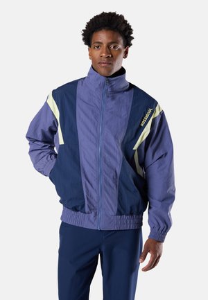 Mann trägt eine blau-lila Zip-Up-Jacke mit gelben Akzenten und „Rossignol“-Branding am Oberarm vor weißem Hintergrund.