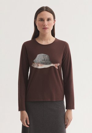Donna con capelli castani lisci che indossa una camicia marrone a maniche lunghe con una grafica di pesce, un cappello a quadri e una gonna con fantasia.
