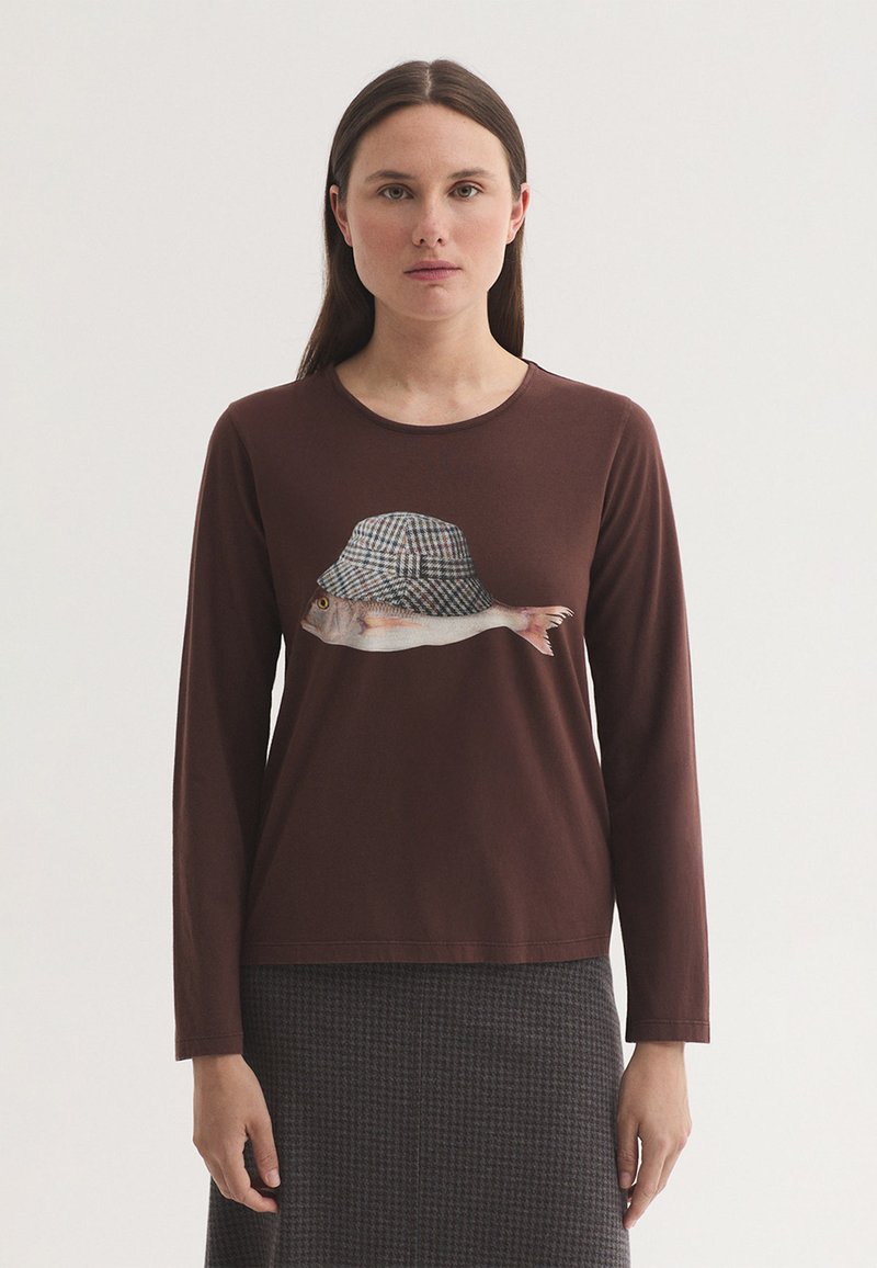 Femme aux cheveux bruns et raides portant une chemise marron à manches longues avec un graphisme de poisson, un chapeau à carreaux et une jupe à motifs.
