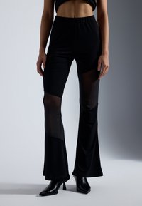 Pantalons noirs évasés en tissu extensible avec des panneaux transparents aux genoux. Taille élastique et jambe fuselée, portés avec des talons noirs.