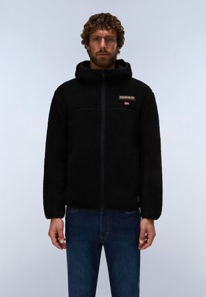 Homme aux cheveux bouclés portant une veste polaire noire zippée avec capuche et un jean bleu, debout devant un fond gris uni.