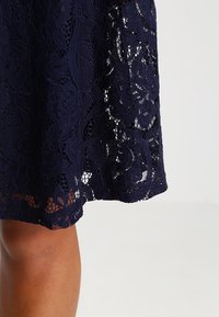 Navy blue lace fabric features floral patterns and open mesh areas. The hem is scalloped, adding texture to the design.