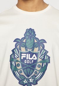 Weißes T-Shirt mit einem lebendigen blauen und grünen FILA Golf-Grafikdesign, das Golfschläger, Bälle und tropische Elemente in einem Schildmotiv zeigt.