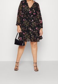 Robe noire à fleurs avec manches longues, décolleté en V, ourlet volanté et motif de fleurs multicolores ; assortie à un sac à main noir et des talons à lanières.