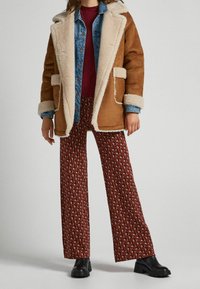 Brun shearlingfoderad rock, blå denimjacka, vinröd skjorta, mönstrade vida byxor i röd och brun, svarta klumpiga stövlar.