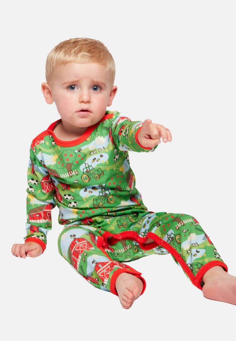 Tout-petit aux cheveux blonds assis, portant un pyjama vert avec des scènes de ferme, pointant devant lui avec une expression préoccupée.