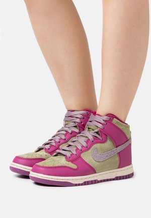 Nogi w różowo-oliwkowych wysokich trampkach z szarymi sznurówkami i teksturowanym logo Nike na białym tle.