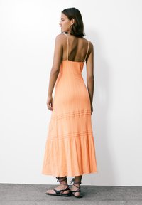 En lang, flydende orange kjole med smalle stropper, tekstureret stof, lagdelt kant og et simpelt design, parret med sorte sandaler.