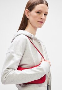 Κόκκινη δερμάτινη τσάντα χεριού με μινιμαλιστικό σχεδιασμό, λεία υφή και μία ενιαία θήκη ώμου, φορεμένη πάνω από ένα ανοιχτό γκρι φούτερ.