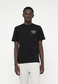 Μαύρη βαμβακερή μπλούζα με στρογγυλή λαιμόκοψη, διακοσμημένη με λευκό κείμενο και γραφικά με αστέρια στο αριστερό στήθος. Εφαρμοστή γραμμή και κοντά μανίκια.