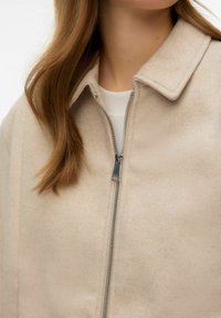 Veste beige à fermeture éclair en tissu texturé, avec un design à col et un détail minimaliste avec une fermeture éclair argentée.