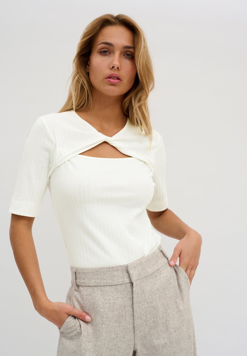 White ribbed top with a knotted front and square cutout, paired with light beige tailored trousers featuring side pockets.