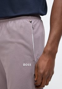 Κοντινό πλάνο ενός ατόμου που φοράει ανοιχτό μωβ φόρμα με ελαστική μέση και λευκό λογότυπο "BOSS" κοντά στην τσέπη, με το χέρι ακουμπισμένο δίπλα.