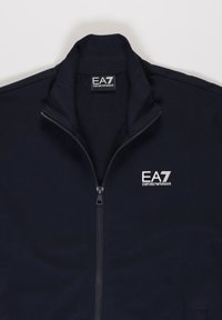 Giacca zip-up blu navy con colletto alto, caratterizzata da un logo bianco EA7 Emporio Armani sul petto sinistro, texture liscia e senza tasche visibili.
