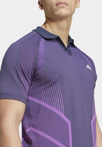 Polo deportivo morado con cuello azul marino, que presenta rayas texturizadas y patrones geométricos en diferentes tonos de morado.