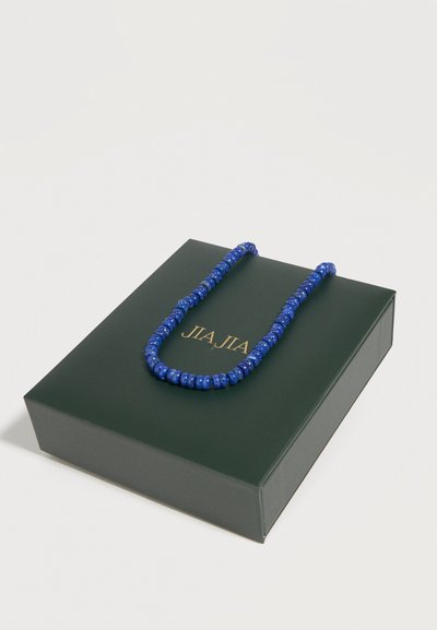 Boîte à bijoux vert foncé avec lettrage doré et un collier de perles bleu posé dessus sur un fond blanc.