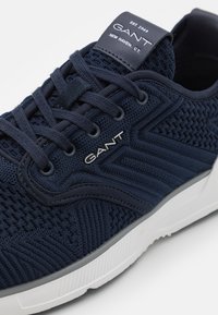 Blue knitted sneaker with a textured surface, featuring a white sole and GANT branding on the tongue. Laces threaded through reinforced eyelets.