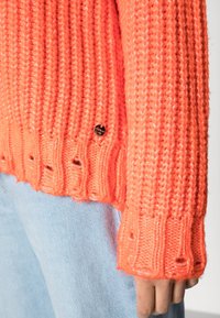 Pull tricoté de couleur corail vif avec texture côtelée, comportant un logo rond en métal et des détails ajourés à l'ourlet et aux poignets.