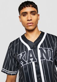 Čierno-biela pruhovaná baseballová dres s krátkymi rukávmi, s predným zapínaním na gombíky a s nápisom "KANI" výrazne zobrazeným.