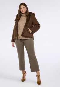 Brązowa kurtka z ociepleniem i sztucznym futrem przy kołnierzu, beżowy sweter dzianinowy z wzorem, brązowe spodnie w kratę i brązowe buty na obcasie.